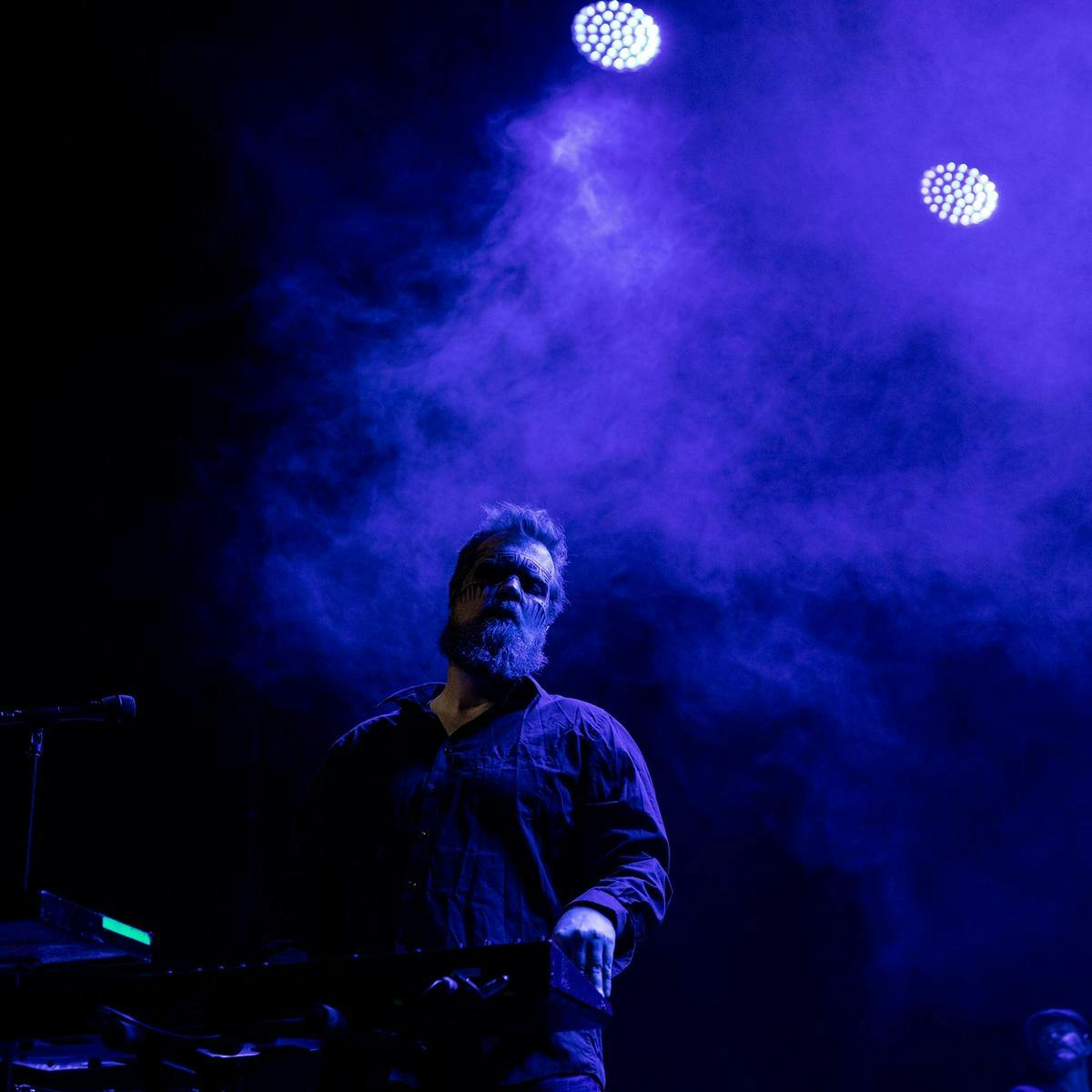 John Grant stands on stage next to his keyboard, purple light and smoke fill the background.