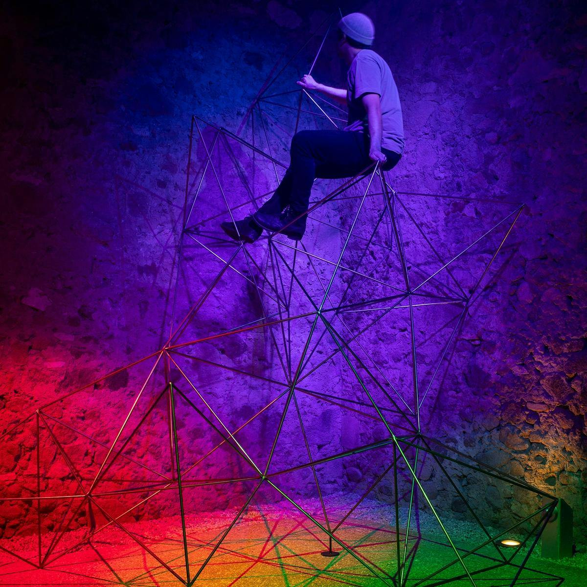 A person sits atop a geometric structure made from metal tubing. Behind the structure is a brick wall lit up by a rainbow of lights.