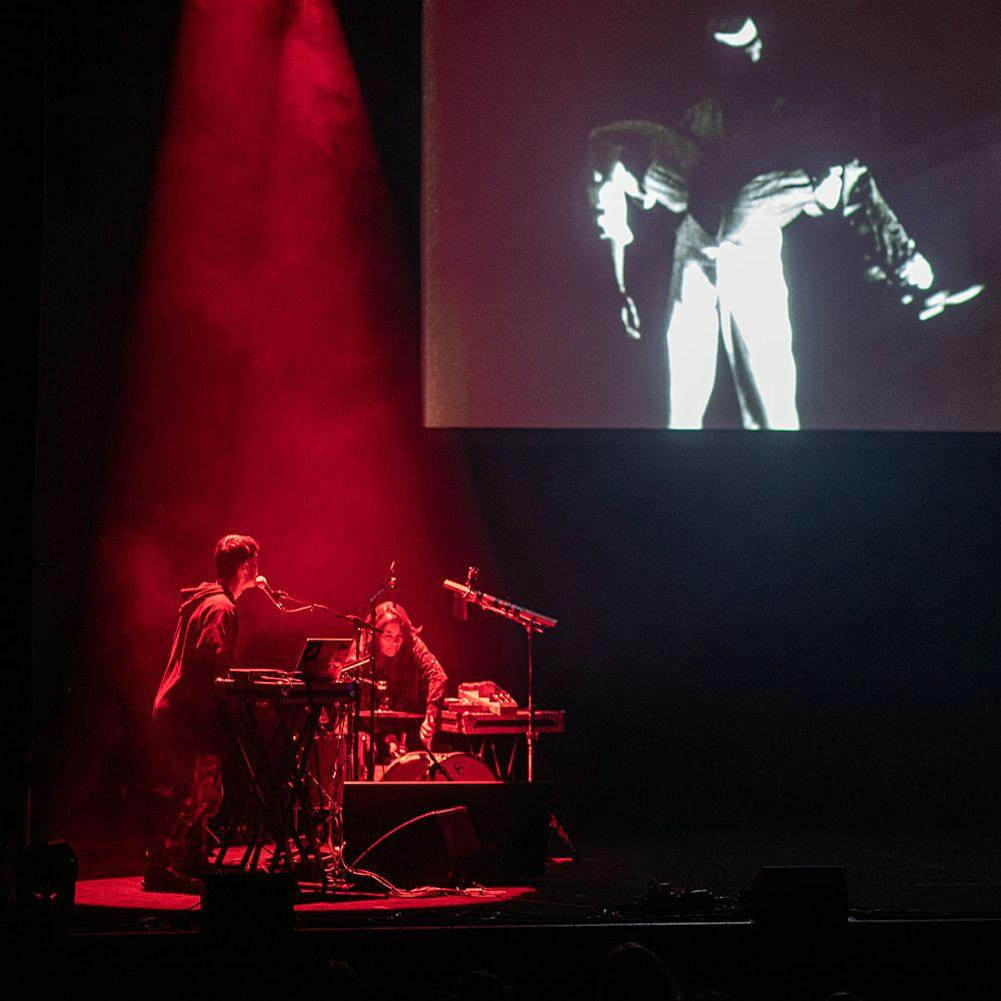 Two performers on stage, behind them is a large screen.