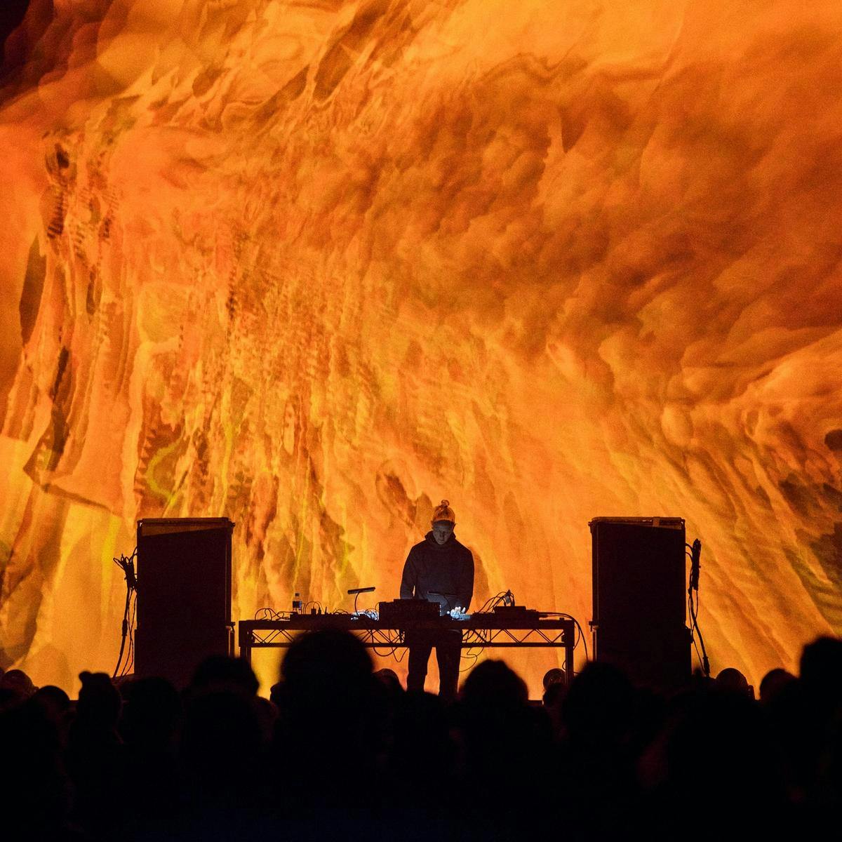 A DJ performing on stage, behind them a large abstract fiery shape is projected onto a screen.