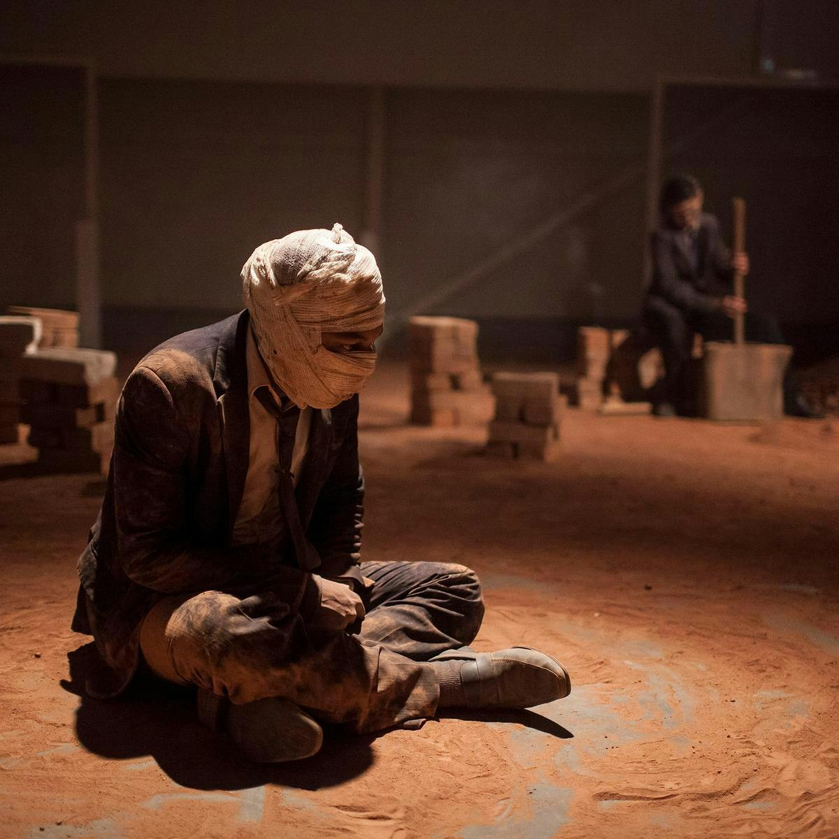 A person sits on the dusty and dirty ground wearing a suit. Their face is hidden by bandages, only their eyes visible. Dirt covers all of their clothes.