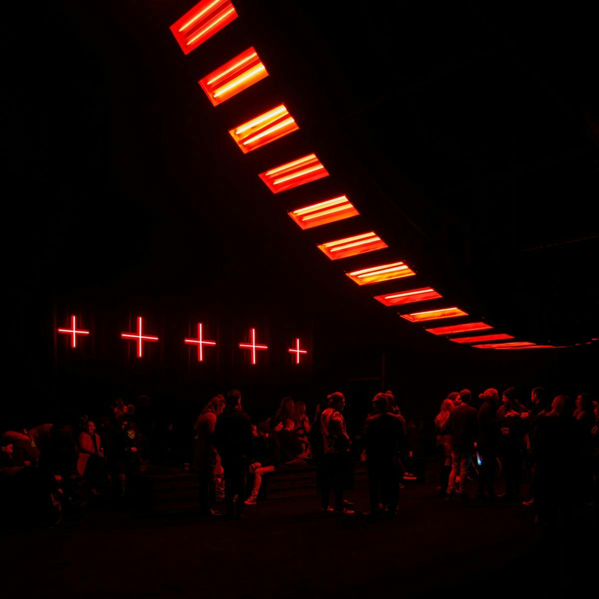 Several people sitting, and standing in a dimly lit room. Several red crosses and red ceiling lights light the room from above.