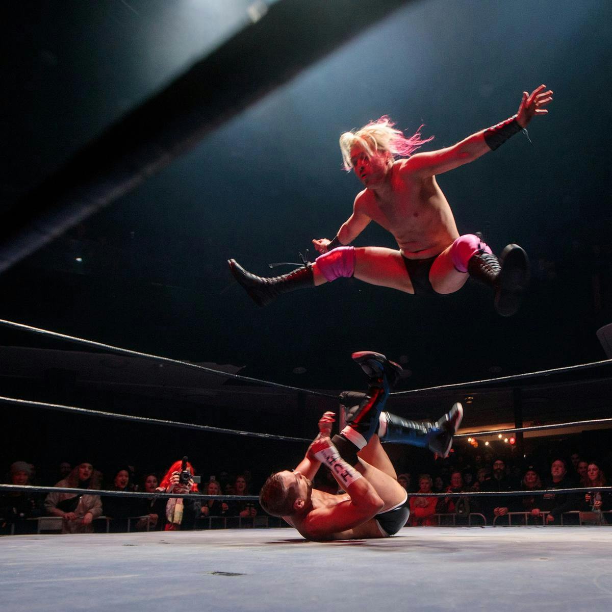 Capture from ground level, a wrestler jumps high into the air, appearing to fall towards another wrestler who is lying on the ground.