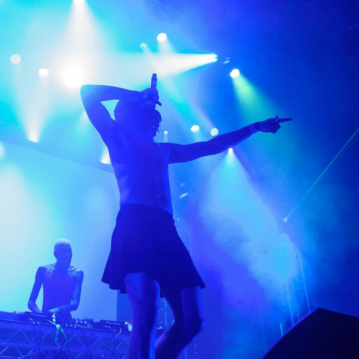 Awash in blue light, a top-less person sings into a microphone whilst pointing out towards the crowd.