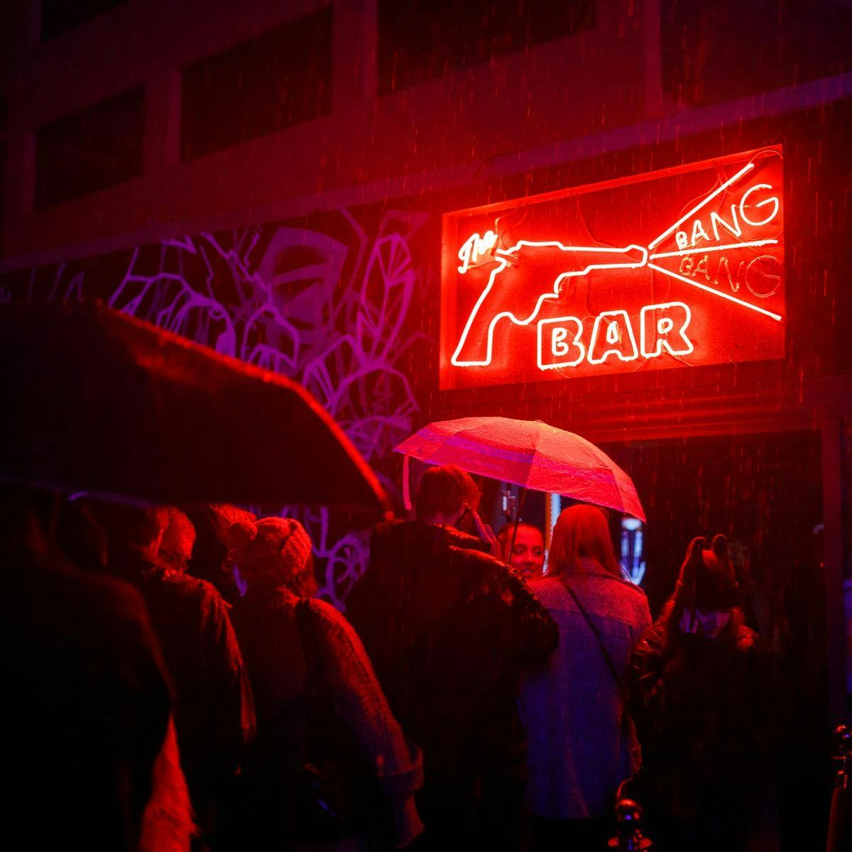 Several people holding umbrellas queue in the rain, waiting to go through a doorway. Above the doorway is a neon sign which reads 'The Bang Bang Bar'.