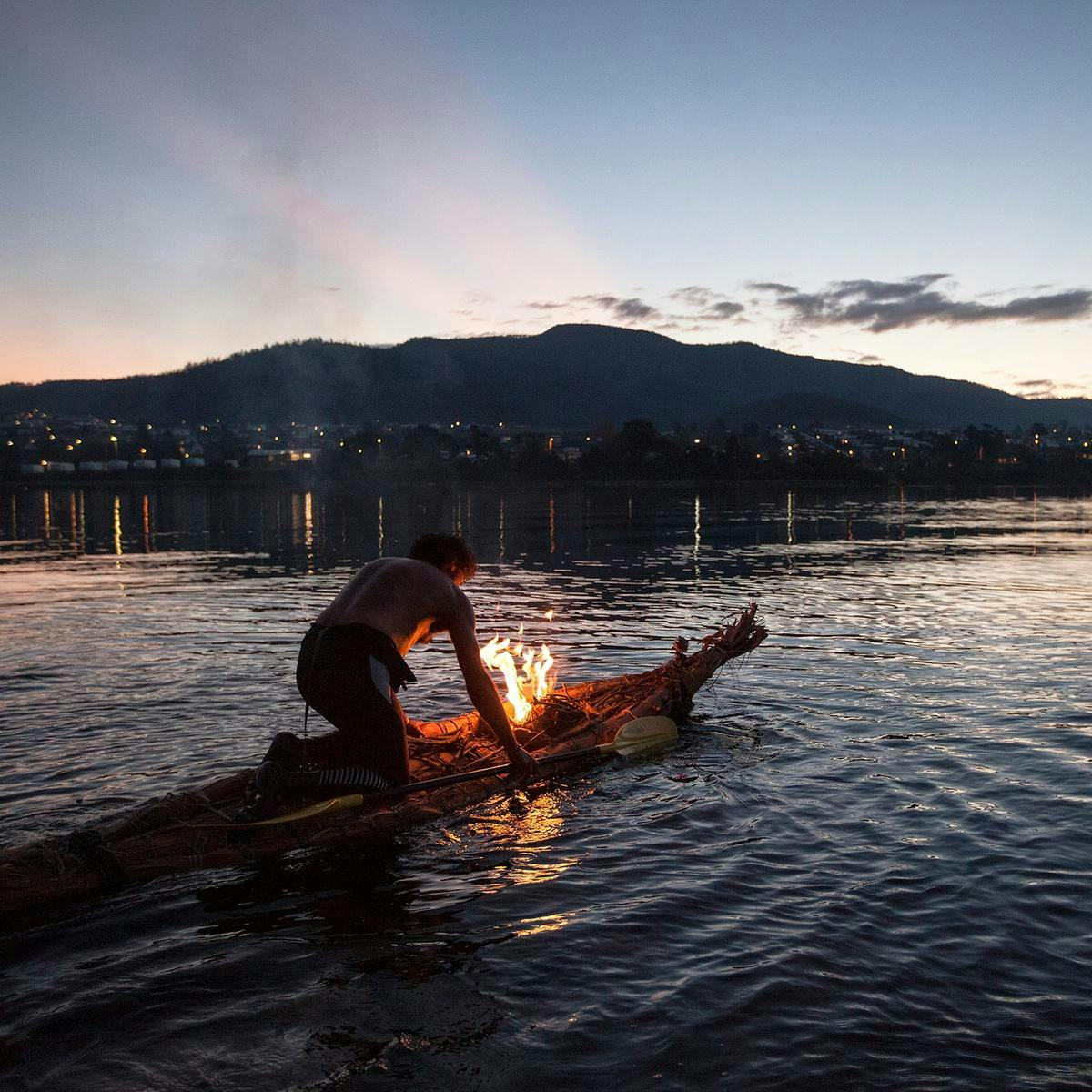 A man kneels down in a traditional Aboriginal bark canoe, tending to a fire as he floats out into a body of water.