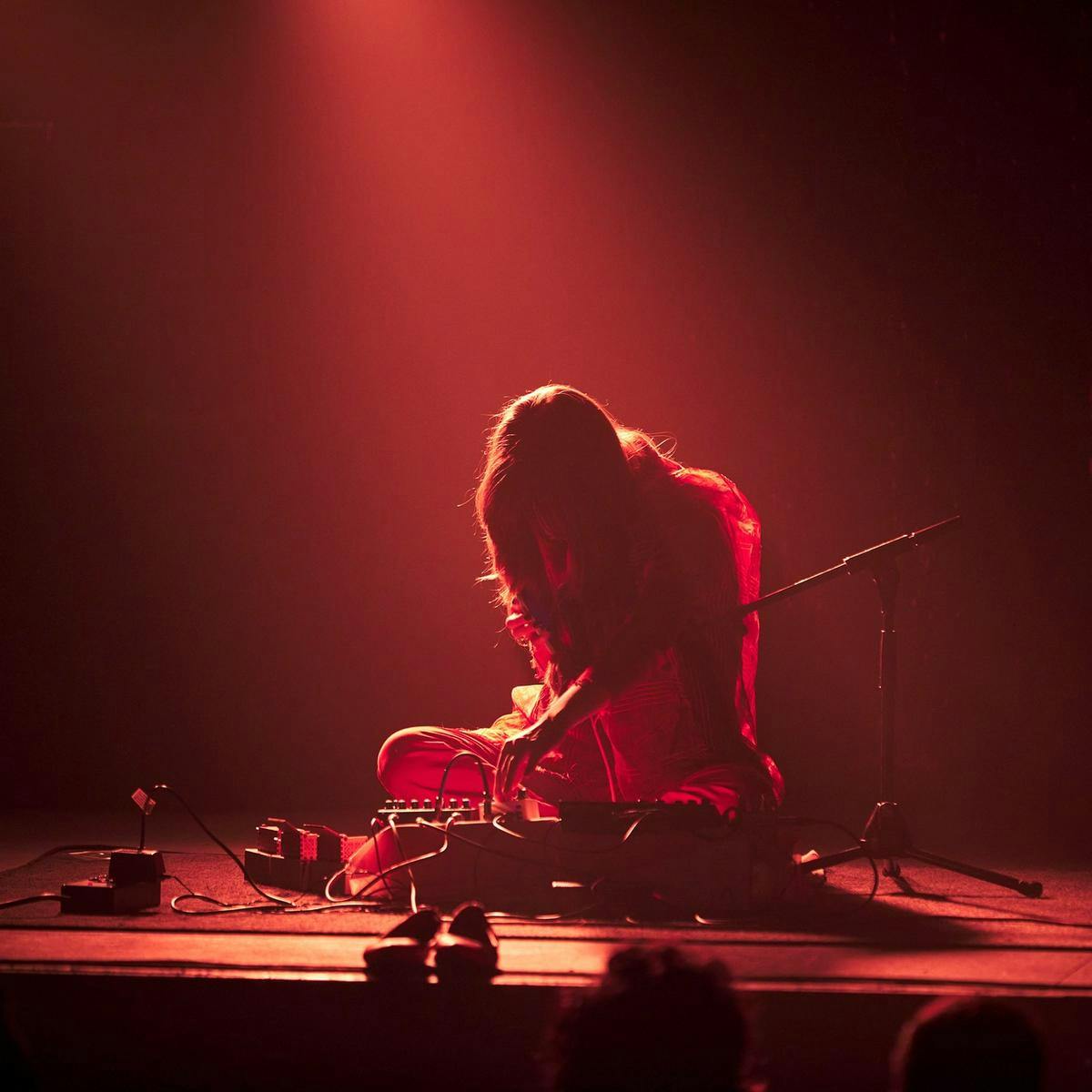 A person sits on stage, adjust a knob on a musical device. Red light provides a hair light to their body.