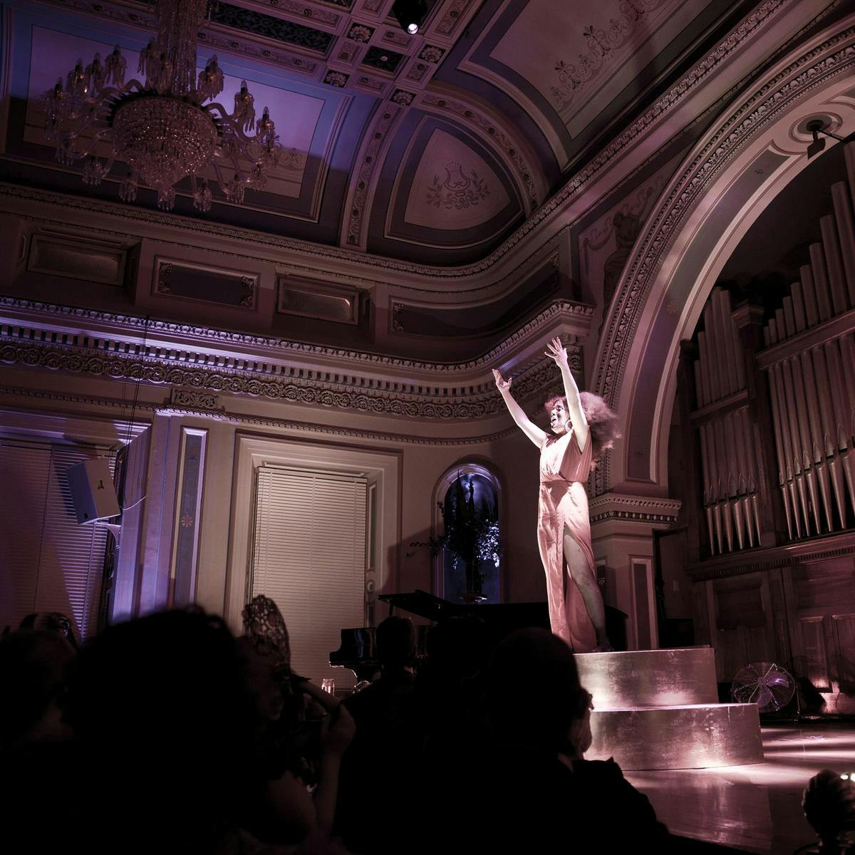 A completely silver painted person stands on stage in front of several people, hands outstretched. Above them an ornate chandelier and vaulted ceiling.