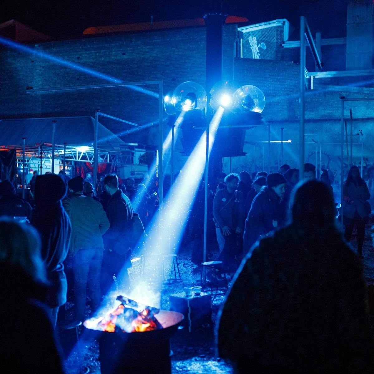 A crowd of people gather outside around fire pits, blue sweeping lights move around the area.