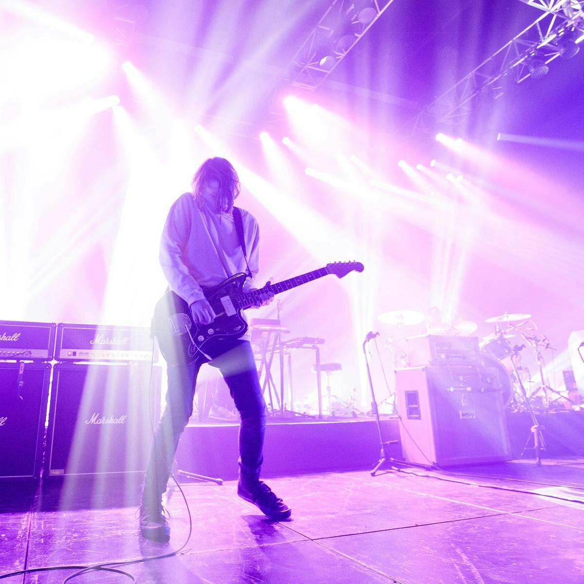 A person performs on stage, several purple lights and speakers are behind them.