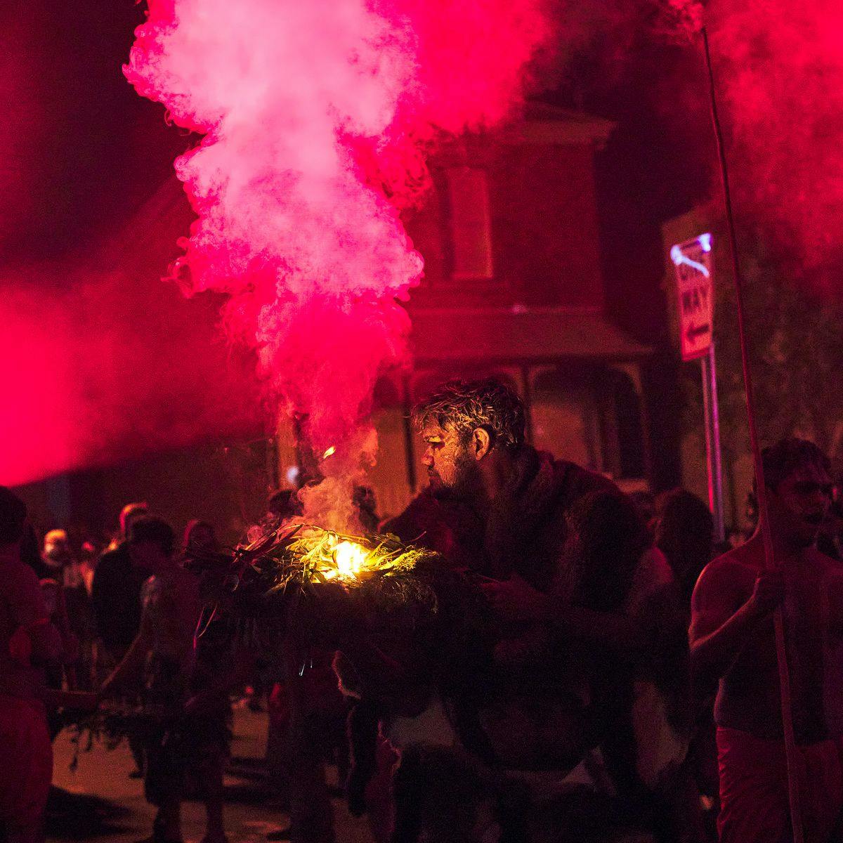 A person stands in the street, wearing several animal furs, whilst holding a smoking bundle of leaves. Behind them are several other people.