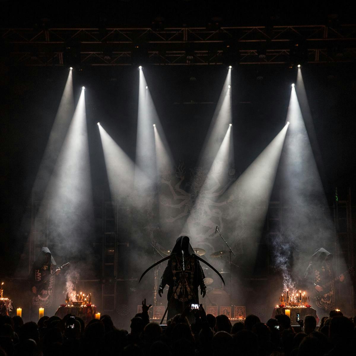 Three hooded figures perform on stage. Many candles burn, with the smoke rising up to meet beams of white light.