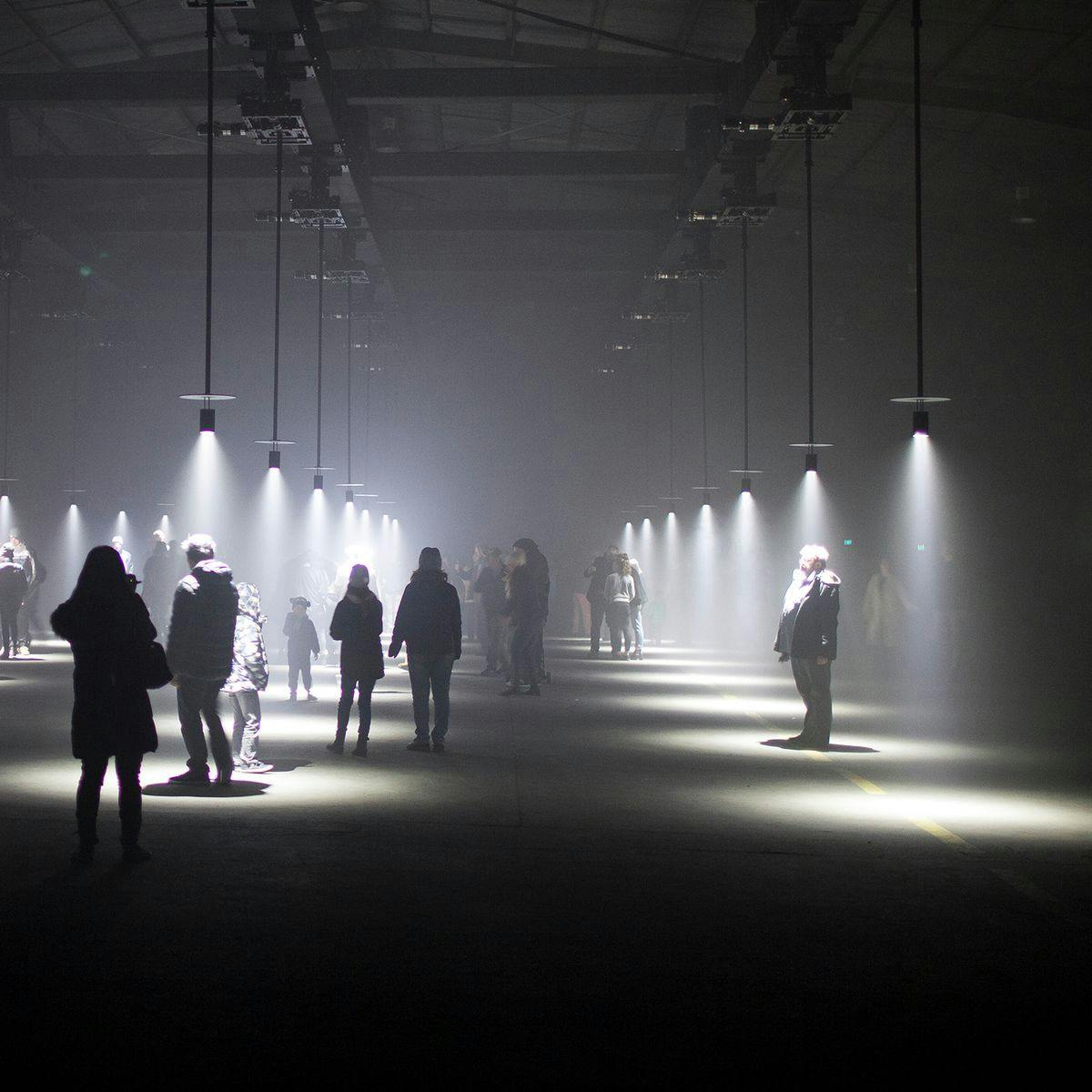 Several people stand and observe the 'Our Time' artwork. Rows of spotlights hang down from a high ceiling.