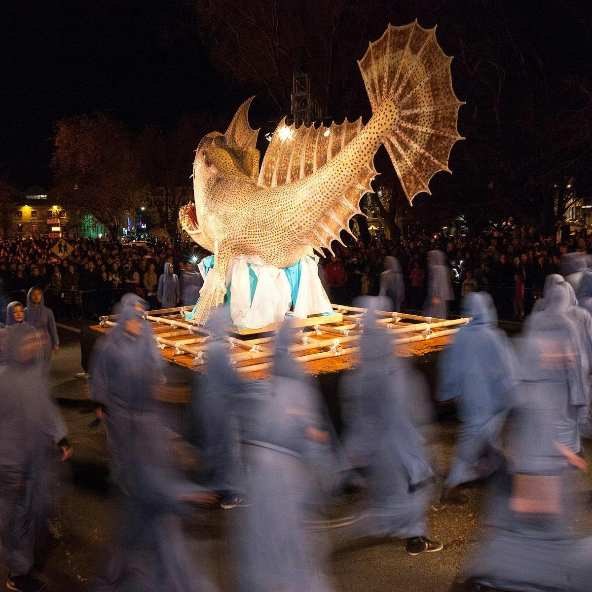 A blurred long exposure image of many hooded figure carrying a fish Ogoh-Ogoh sculpture.