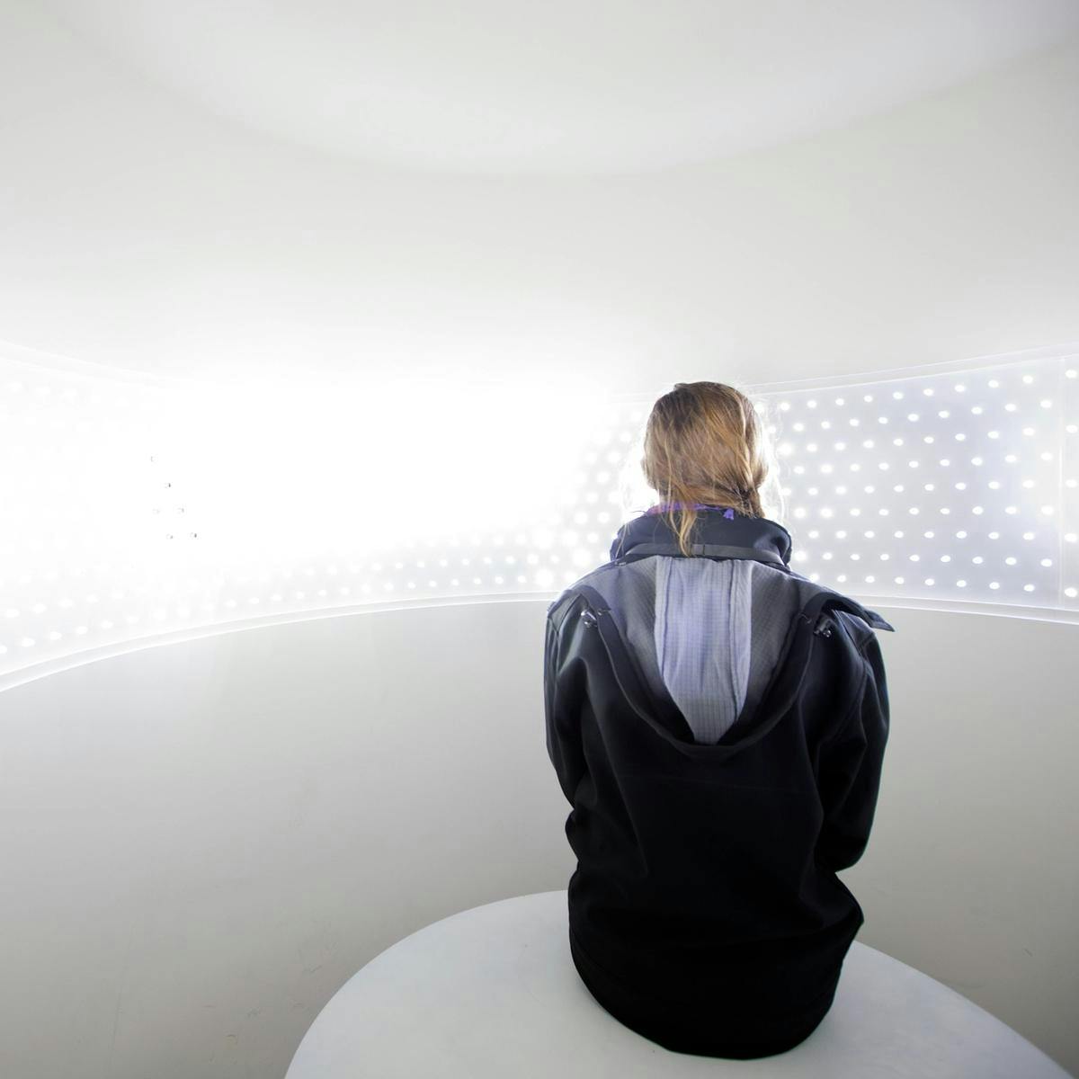 Person sitting with their back to camera in a white room, with hundreds of little light wrapping around the room.