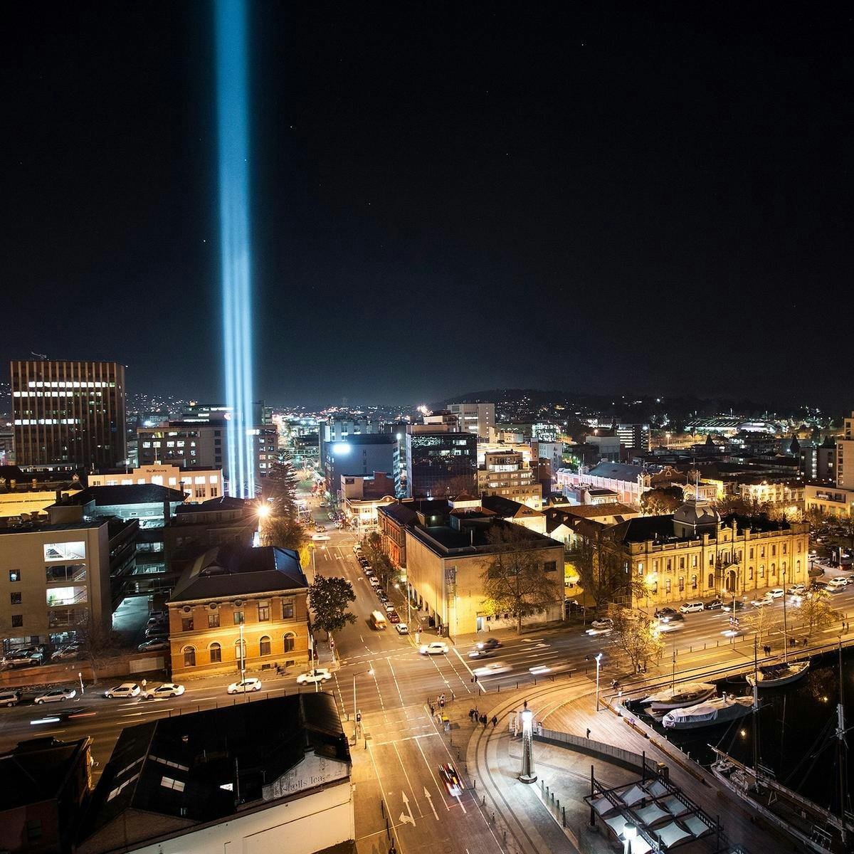 A night-time Hobart cityscape consisting of many buildings and cars, a blue light beams into the sky.