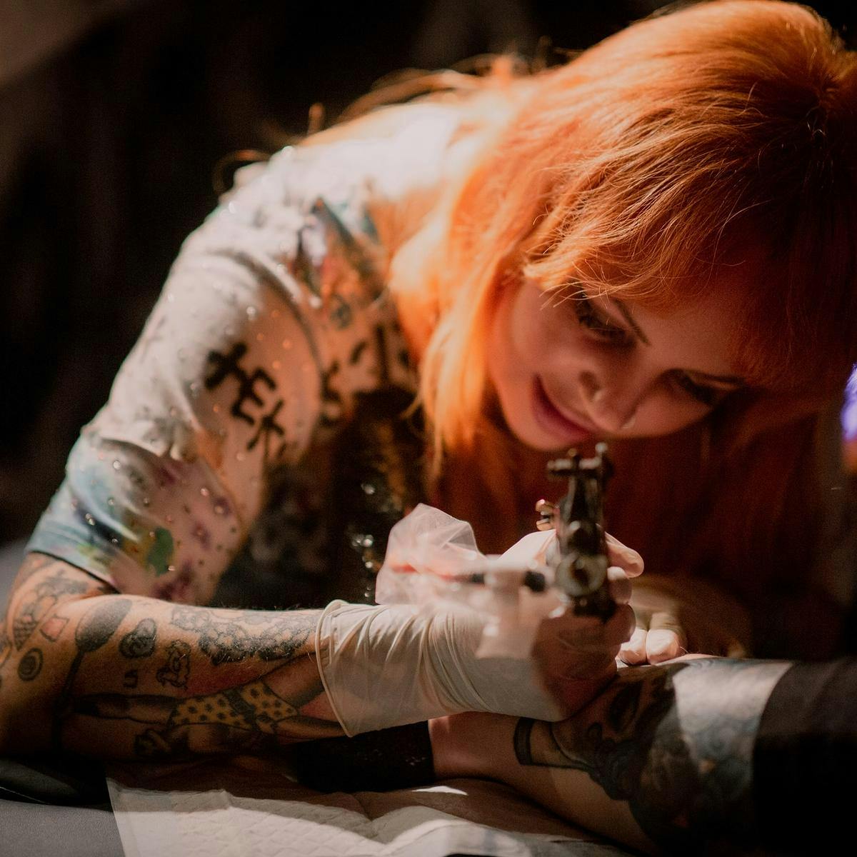 An orange-haired person wearing surgical gloves tattoos another person's calf.