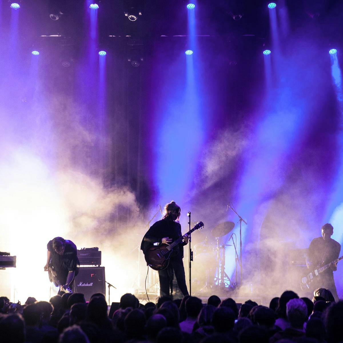 A wide shot of Chelsea Wolfe and their band performing on stage. Blue beams of light and heavy fog surrounds them.