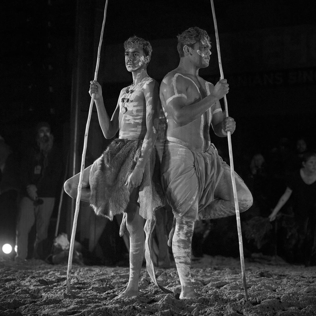 Two people wearing body paint stand back-to-back, holding long spears.