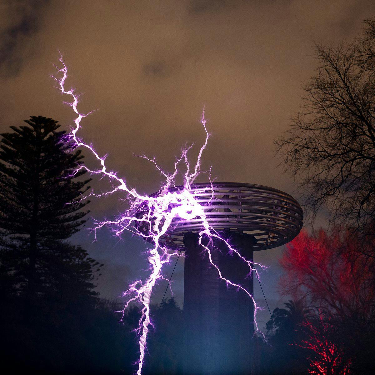 A large tower tesla coil arcs purple lightning into the night's sky. Several trees, and dark clouds adorn the background.