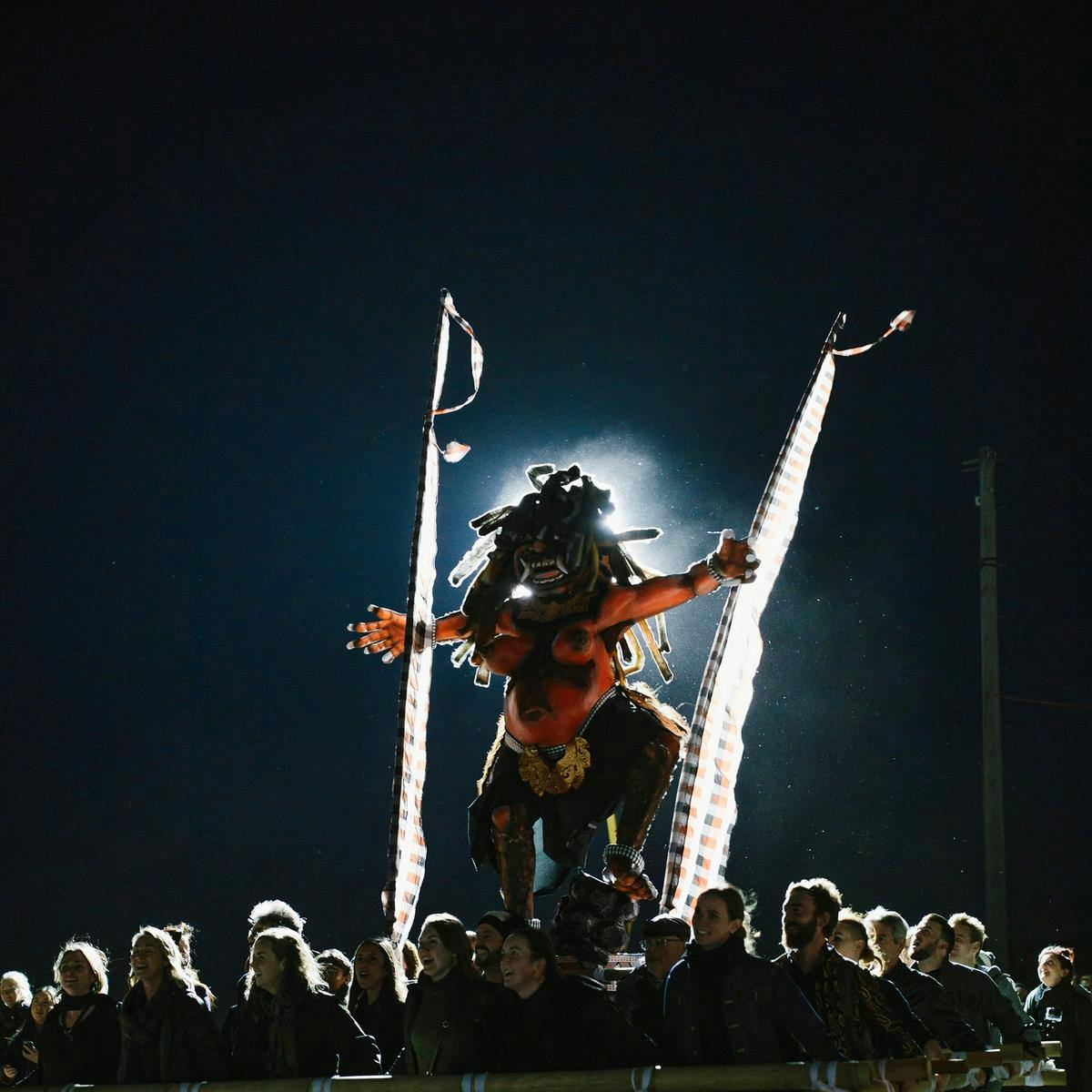 Several smiling people carry a large platform, atop the platform a Balinese human-like sculpture stands.