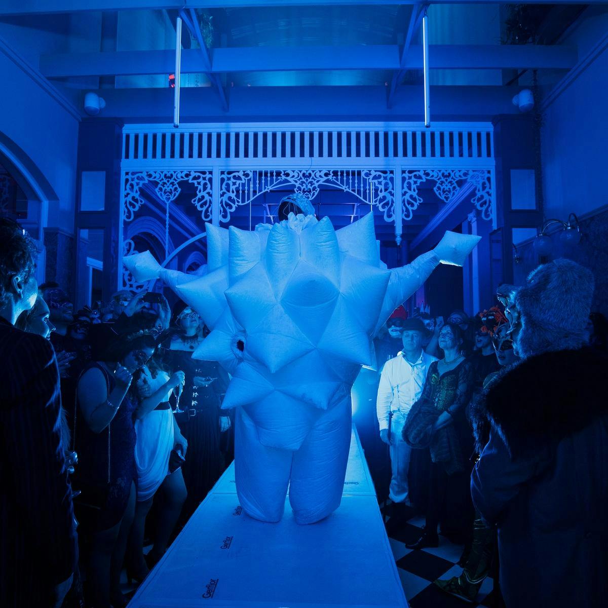 Several costumed people watch a performer walk along a catwalk style stage. The performer is wearing a plush spiked outfit that obscures much of their body.