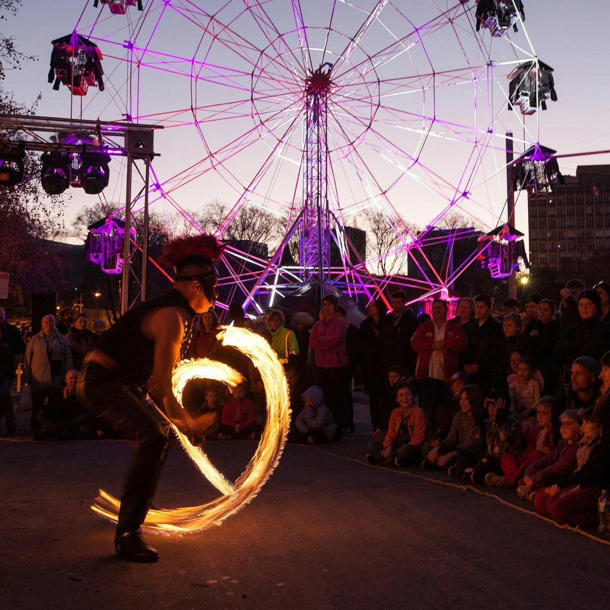 A fire-twirler performs to a crowd at Winter Feast, in the background is a ferris-wheel.