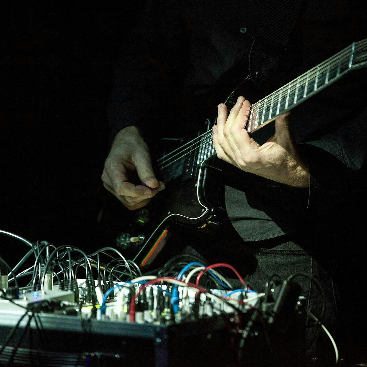 A close up of Marco Fusinato's hands performing a guitar on-stage. In the foreground is a heap of wires, plugged into a MIDI.