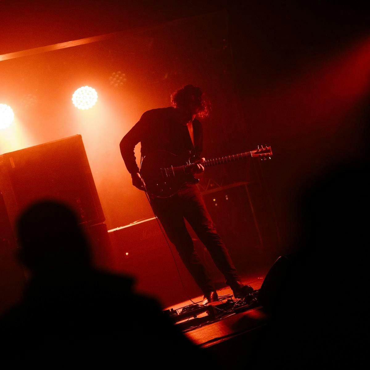 A person performs guitar on stage, they are looking down to their foot pedal.