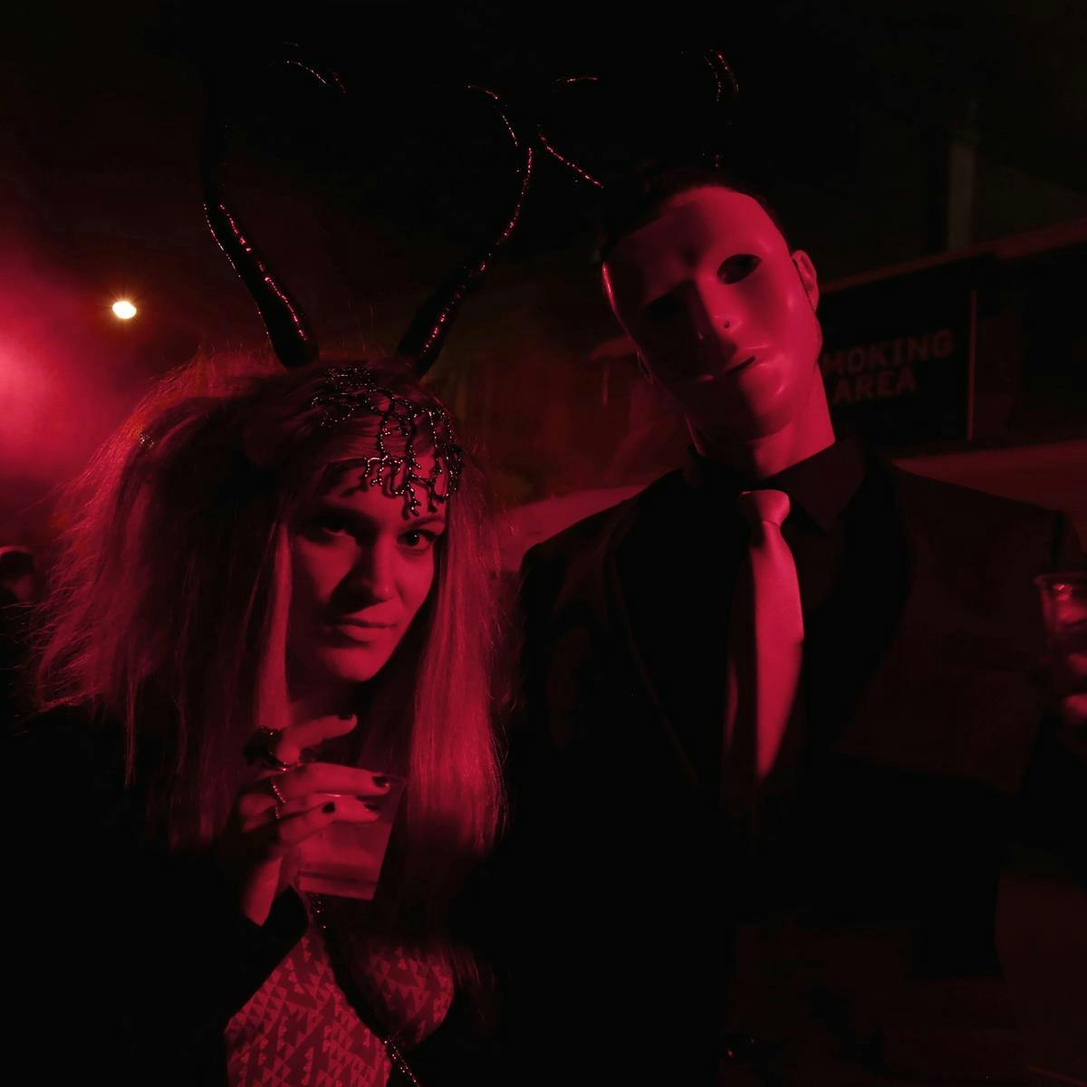 Two costumed people hold drinks up, their faces and bodies lit up with red lights.