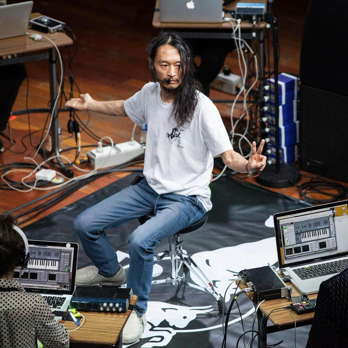 Several people sit at music production workstations in a circular formation, in the centre of the circle a person sits on a stool.