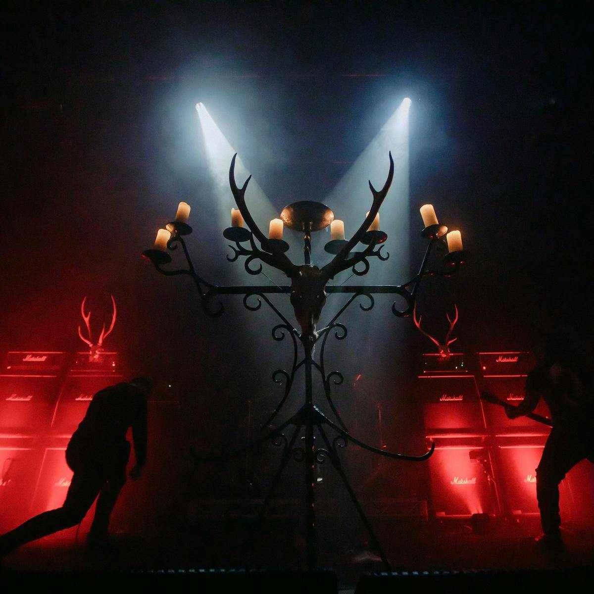 A large candelabra adorned with an animal skull sits centre stage, in the background there are musicians playing guitar and several large speakers.