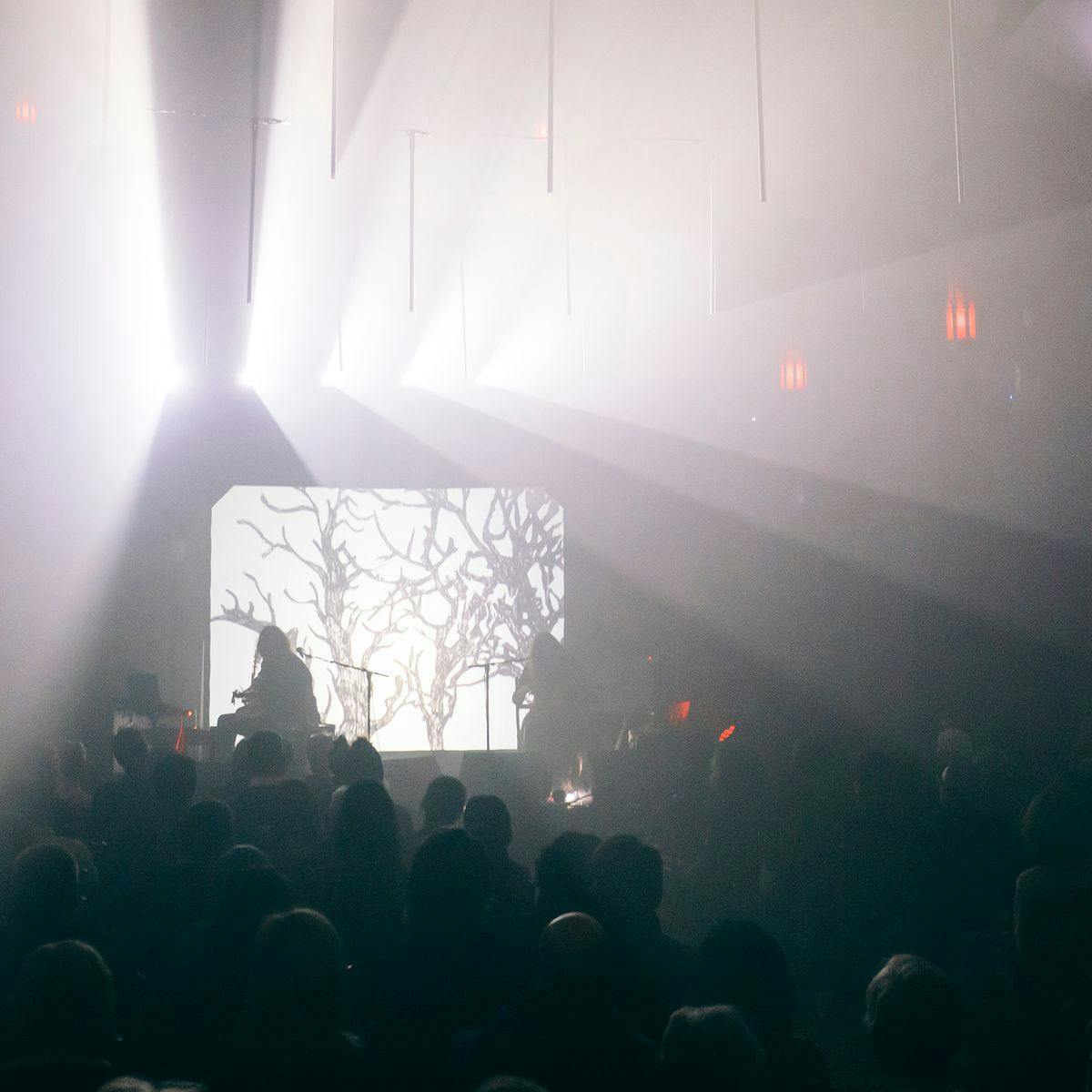 Two performers are on stage infront of a seated crowd. White lights shine from the roof of the building, a small church.