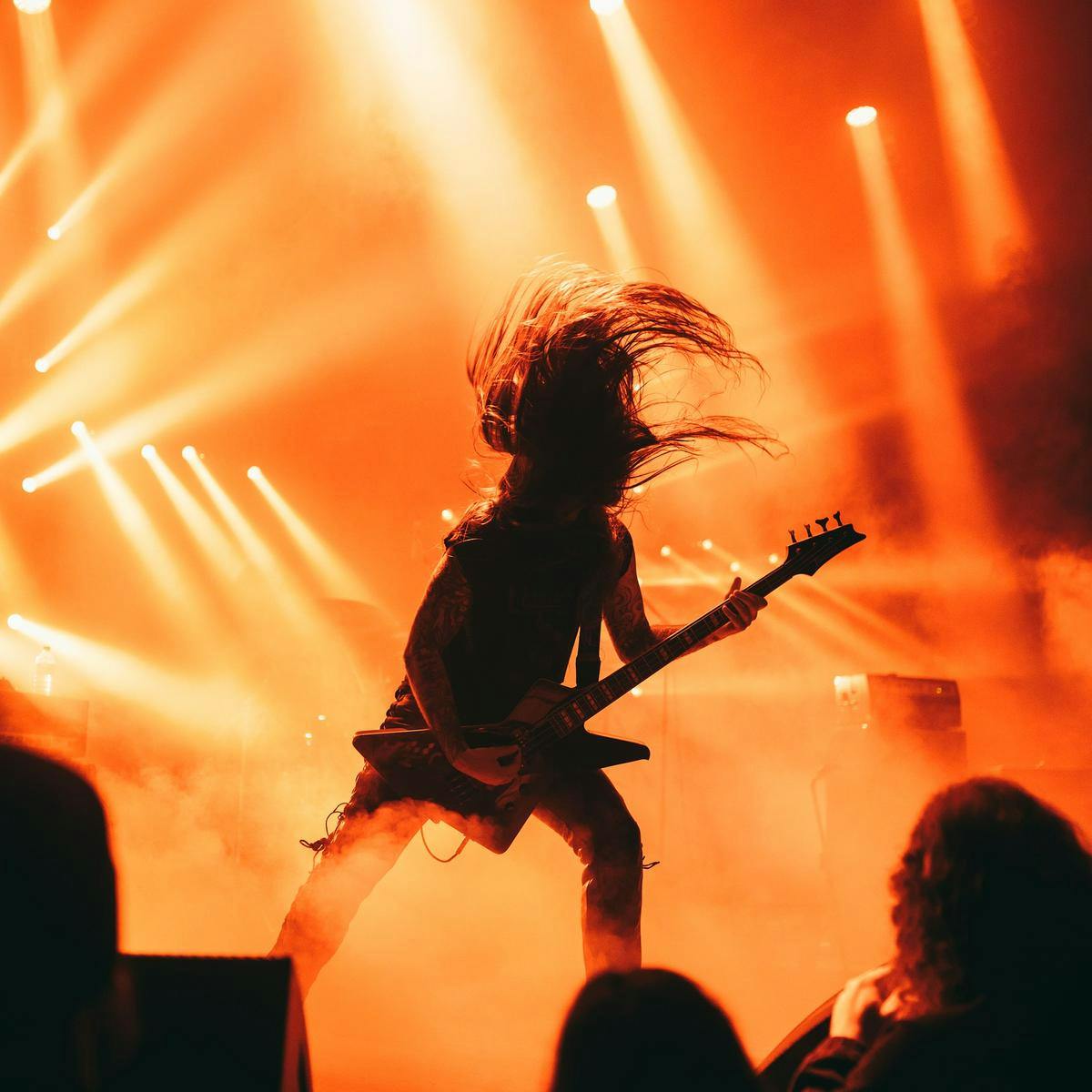 A silhouetted person plays guitar on stage, their hair in motion as they head-bang.