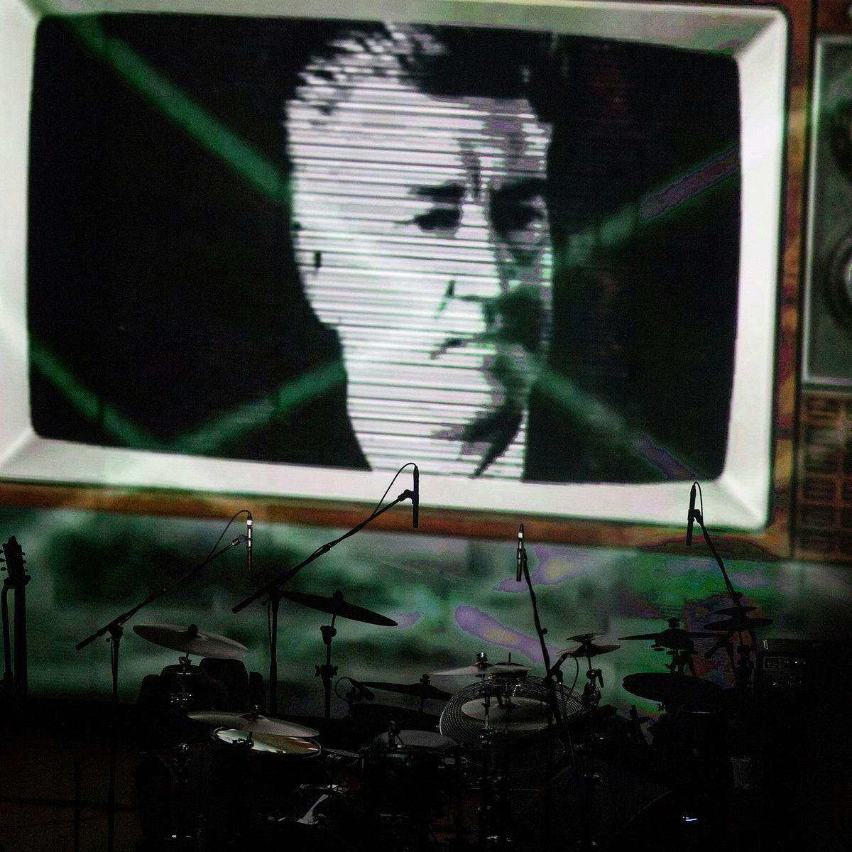 A stage setup consisting of a large drumkit, behind the drums a large projected image of David Lynch within and old-school TV.