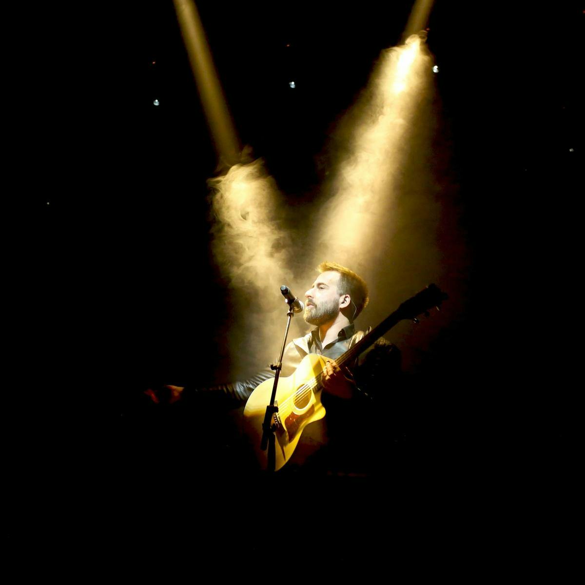 A person performs with a guitar and microphone, two golden light beams highlight them.