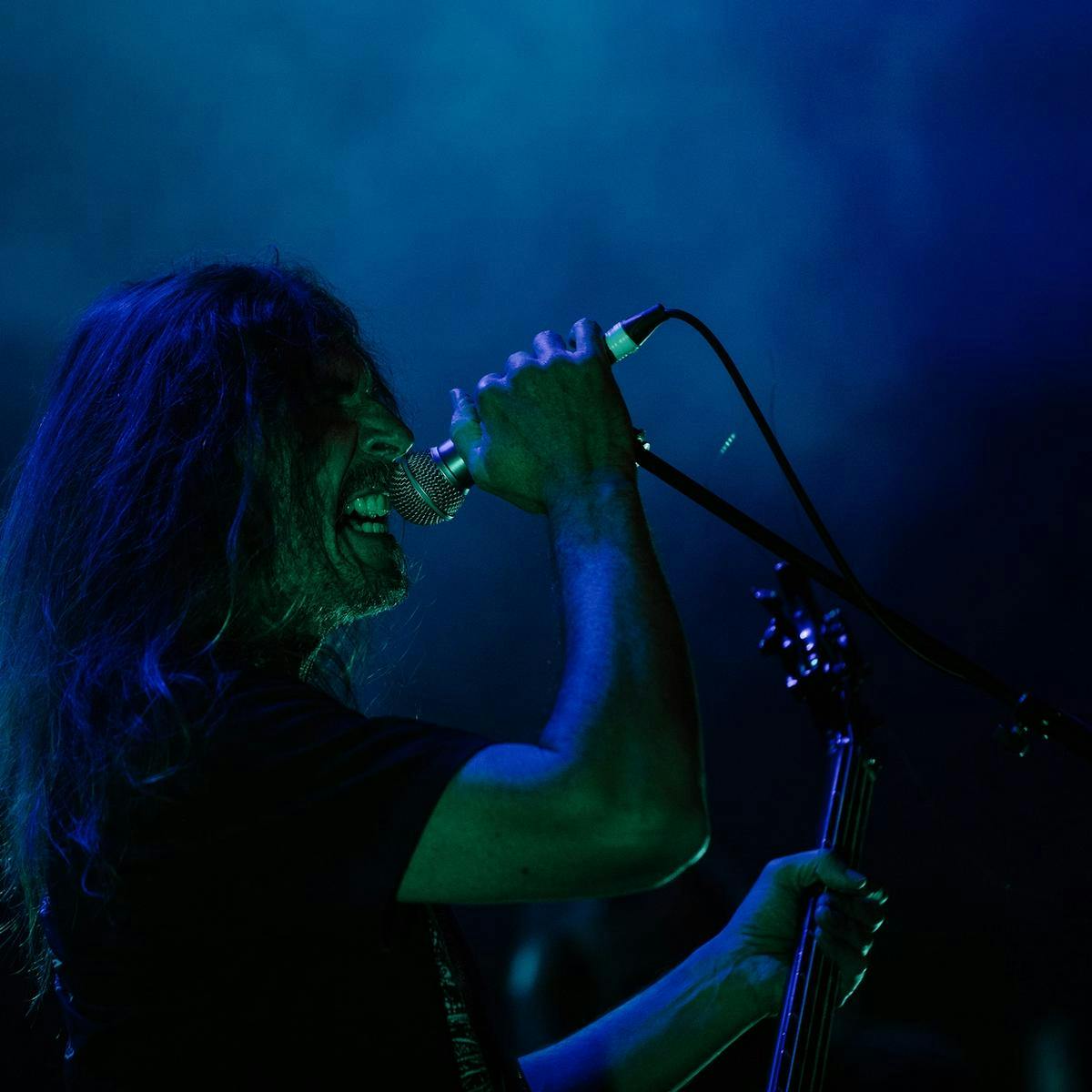 A person performs into a microphone, whilst holding a guitar. They have long hair which covers some of their face.