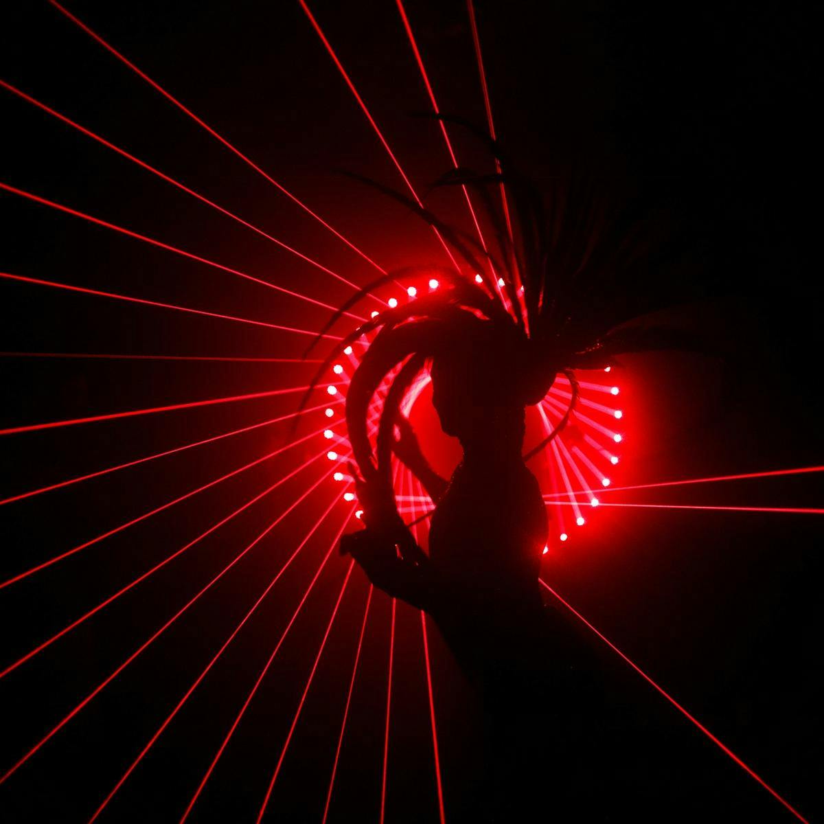 A silhouetted person wears an ornate feathered headpiece. A ring of several red lasers pierce through the darkness.